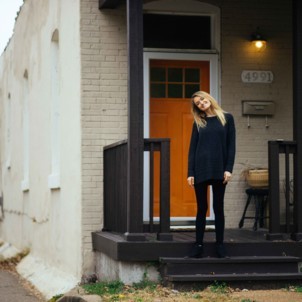 A house with a red door and a happy woman standing outside in a sweatshirt